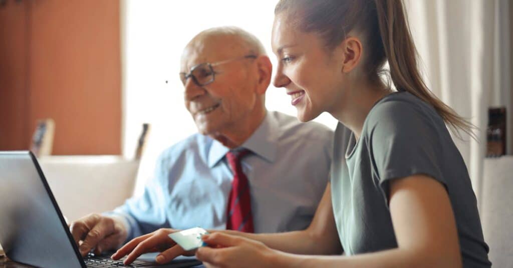 young woman in casual clothes helping senior man in formal shirt with paying credit card in internet- Thom Tax